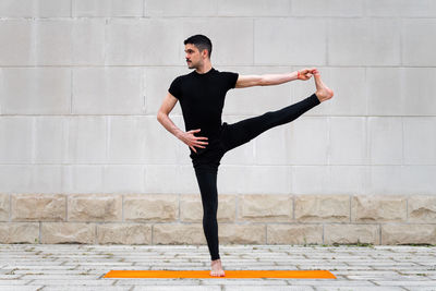 Full length of man doing yoga while standing on mat against wall