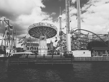 Amusement park by lake against cloudy sky