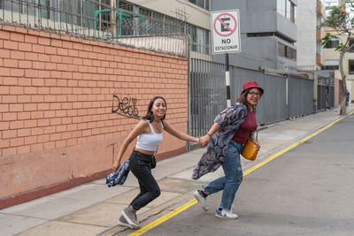 Full body of positive peruvian female friends holding hands and running while crossing road on street with building in city