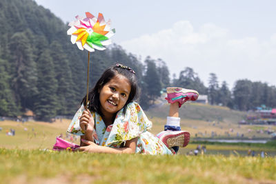 A cheerful smiling indian girl child lying on green grass field looks  with a spinning wheel toy 