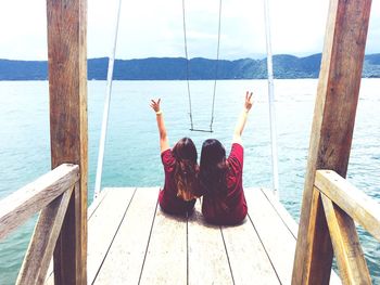 Rear view of friends with arms raised while sitting on pier at sea