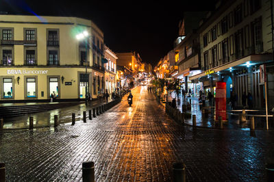 Illuminated street amidst buildings in city during rainy season