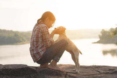 Side view of girl with dog sitting by lake against sky