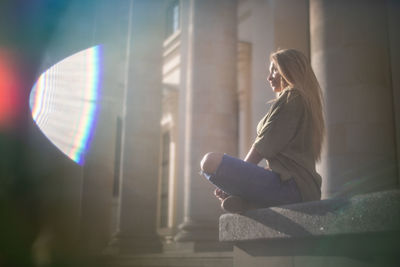 Side view of woman sitting by column during sunny day