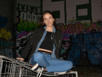 Portrait of young woman sitting on cart against graffiti wall at night