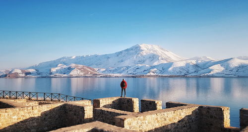 Rear view of man standing on stone wall by lake during winter