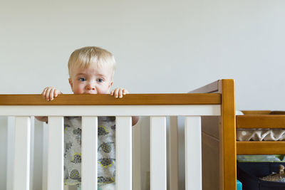 Cute baby boy biting wooden railing while standing in crib at home