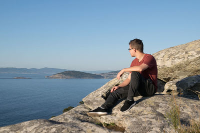 Young man sitting on rock against sky