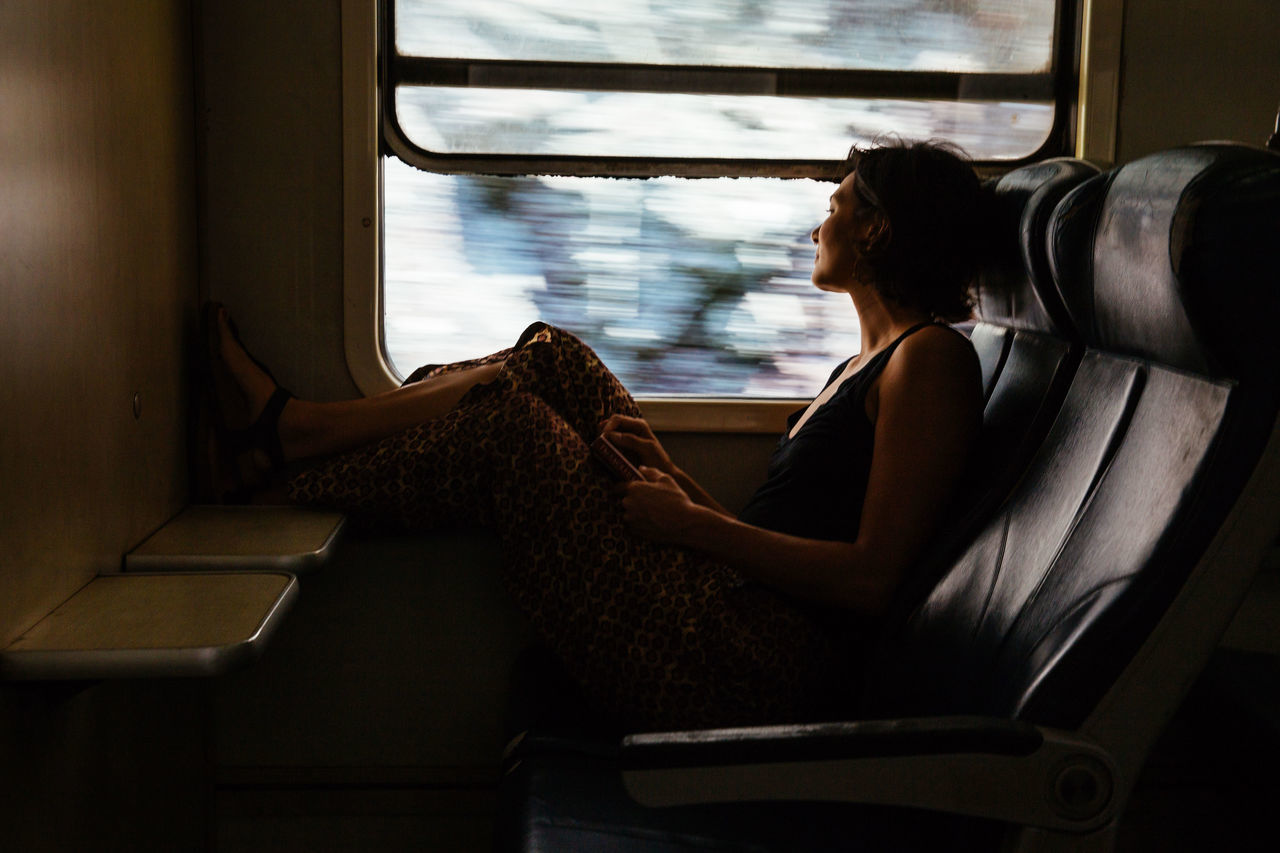 SIDE VIEW OF WOMAN SITTING ON WINDOW AT HOME