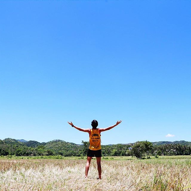 full length, lifestyles, leisure activity, casual clothing, clear sky, copy space, grass, person, standing, young adult, blue, childhood, field, arms outstretched, landscape, elementary age, rear view, tranquil scene