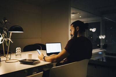 Male entrepreneur using smart phone while sitting with laptop at illuminated desk working late in office