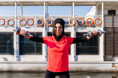 Attractive young woman is doing exercises with dumbbells in front of a boat in the harbor