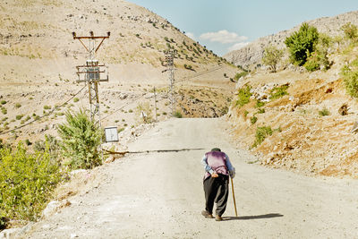 Rear view of senior man walking with stick on dirt road