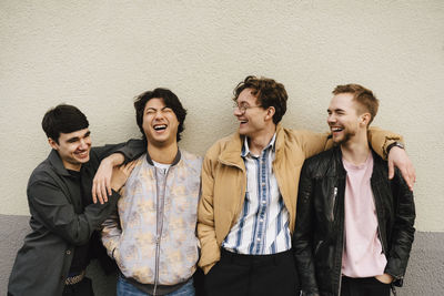 Cheerful male friends laughing while standing against beige wall