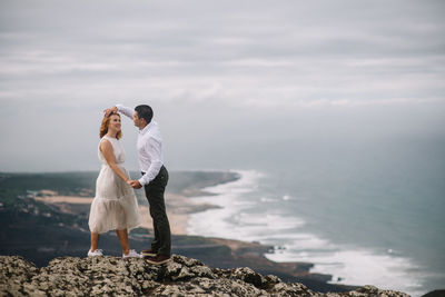 Romantic couple standing on cliff by sea against sky