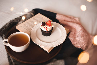 Cup of green tea with tasty cupcake and open book on knitted sweater in bed closeup. good morning.