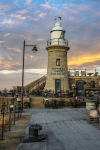 Lighthouse by buildings against sky at sunset