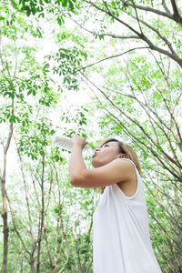 Woman drinking water from bottle while standing against trees in forest