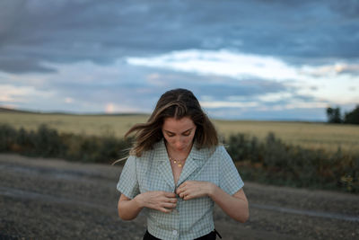 Trendy young female buttoning up shirt on roadway against field under cloudy sky in twilight