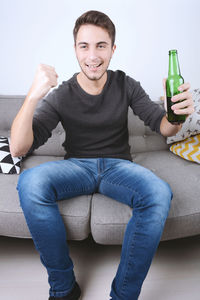 Smiling young man watching tv while holding drink on sofa at home