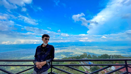 Young man standing on railing against blue sky