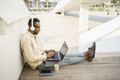 Freelancer using laptop wearing headphones sitting by wall