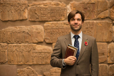 Portrait of smiling man holding books while standing against wall