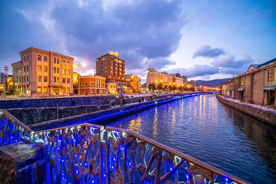 Illuminated bridge over river by buildings against sky at dusk