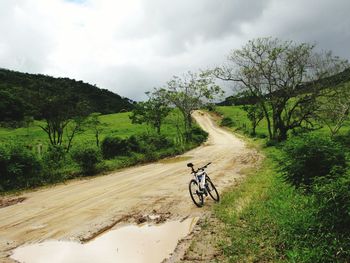 Bicycle parked by puddle in dirt road against cloudy sky