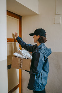 Side view of female delivery person holding parcels and knocking door in building