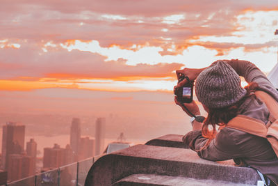 Rear view of woman photographing with camera while standing at beach against sky during sunset