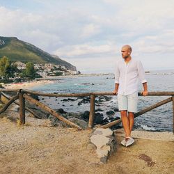 Young man looking away while standing at beach against cloudy sky