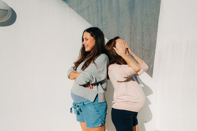 Two pregnant women standing against wall during sunny summer day