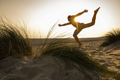 Back lit shirtless young man practicing yoga amidst plants at beach against clear sky during sunset