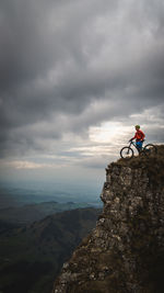 Man with bicycle standing on cliff against cloudy sky