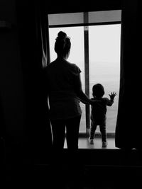 Rear view of mother and daughter looking through window at home