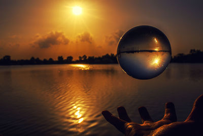 Cropped hand levitating crystal ball by lake during sunset