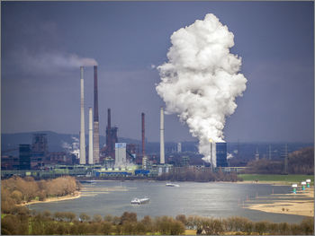 Smoke emitting from chimney by river against sky