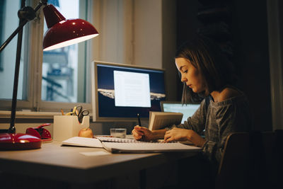 Side view of girl writing while using computer at illuminated desk