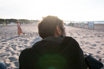 Rear view of man sitting at sandy beach on sunny day