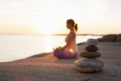 Side view of woman sitting in lotus position on lakeshore with focus on stack of stones