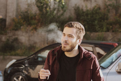Young man looking away while smoking cigarette against cars