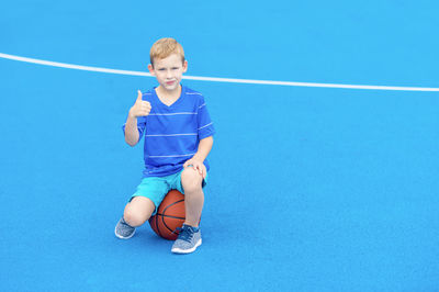 Portrait of boy gesturing thumbs up while sitting on basketball at court