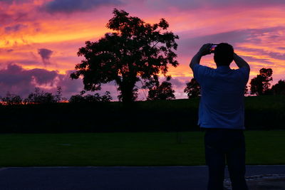 Rear view of man photographing while standing on field against sky during sunset