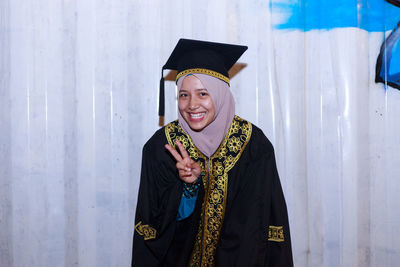 Portrait of smiling young woman wearing mortar board while standing against wall