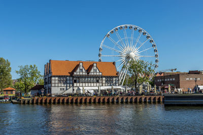 Ferris wheel by river and buildings against clear blue sky