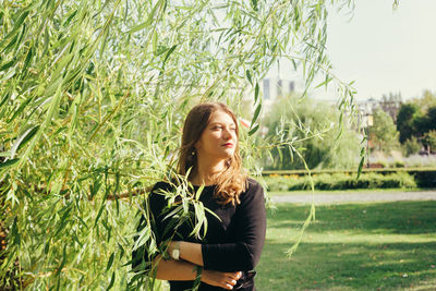 Mid adult woman with arms crossed looking away while standing by plants in park