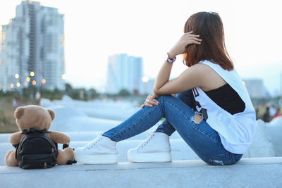 Side view of woman sitting on railing in city against clear sky during sunset