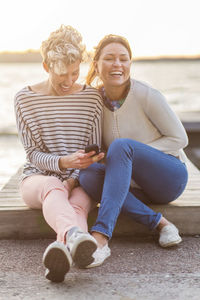 Portrait of happy woman sitting with female friend using mobile phone on pier