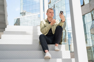 Young man sitting on the stairs and talking sign language via video link on smartphone outdoors